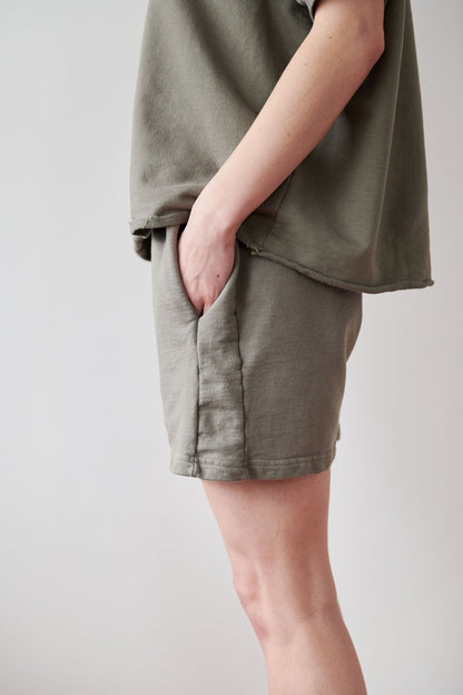 A person wearing a matching olive green shirt and high-rise, elastic waist Wol Hide Cotton Easy Shorts in Fir, made in the USA, stands with their right hand in their shorts pocket. Only the torso and upper legs are visible against a neutral background.