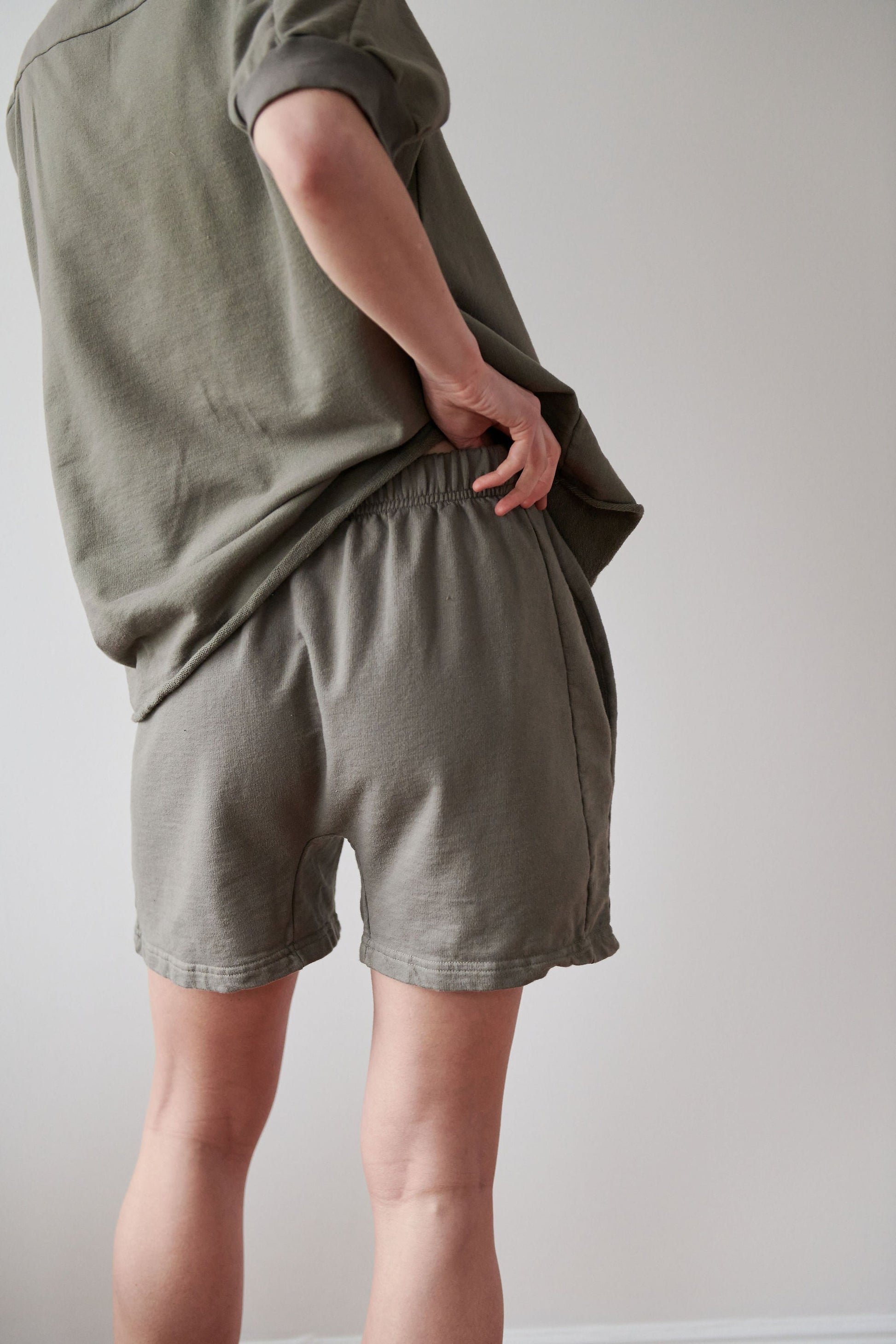 A person standing indoors with their back turned, wearing an olive green long-sleeve shirt and matching Cotton Easy Shorts in Fir by Wol Hide. The individual's right hand is placed on their waist, highlighting the high-rise elastic waistband of the made-in-USA shorts. The background is plain off-white.