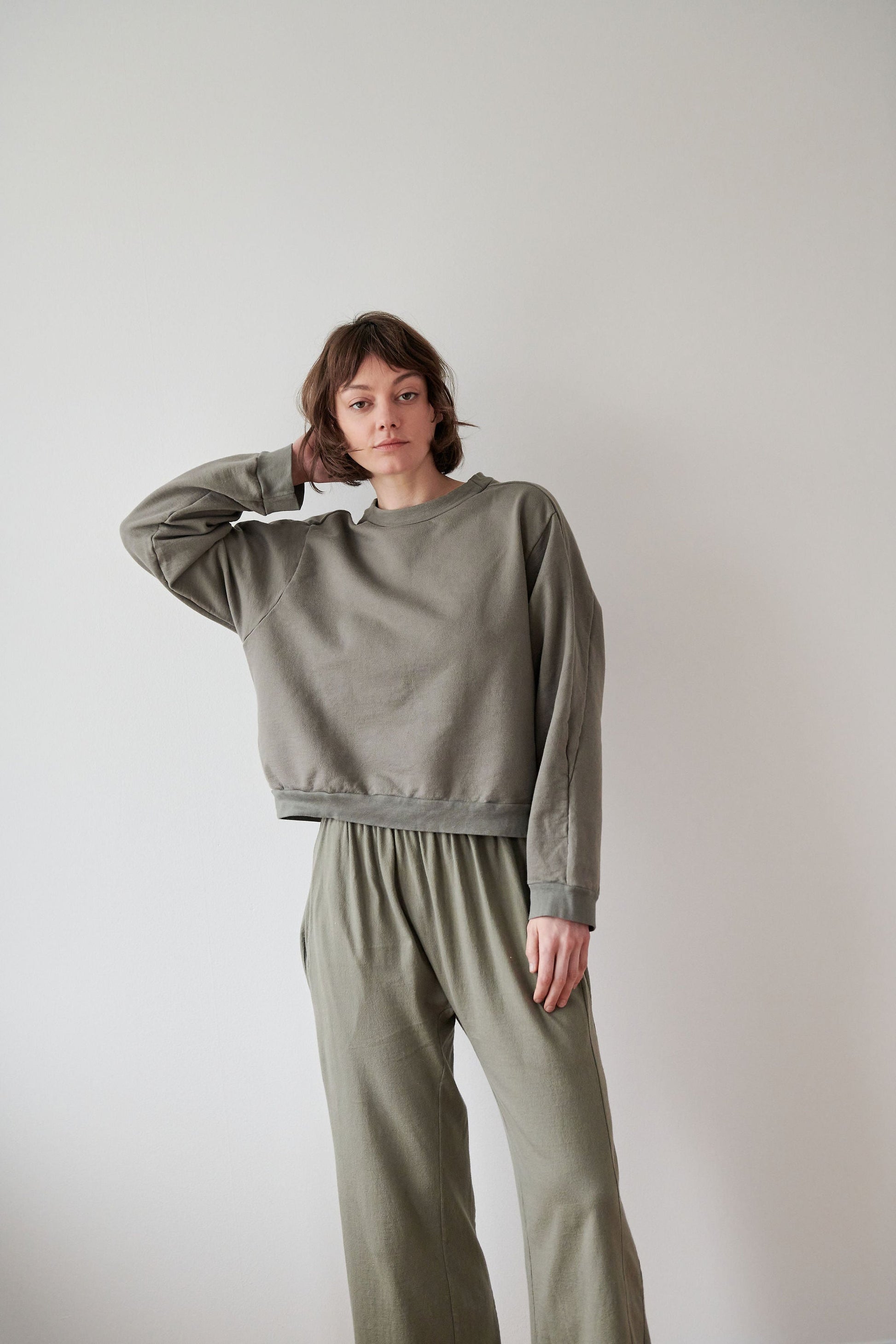 A person with short hair stands against a plain white background, wearing a matching grayish-green Cotton French Terry Easy Summer Sweatshirt in Fir and sweatpants from Wol Hide. Their right hand is raised, touching the back of their head, while the left arm hangs by their side. They have a neutral expression.