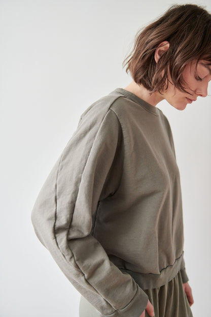 A person with short, tousled brown hair is standing sideways and slightly bowed. They are wearing an olive-green, long-sleeved Cotton French Terry Easy Summer Sweatshirt in Fir by Wol Hide with a matching bottom, set against a plain white background.