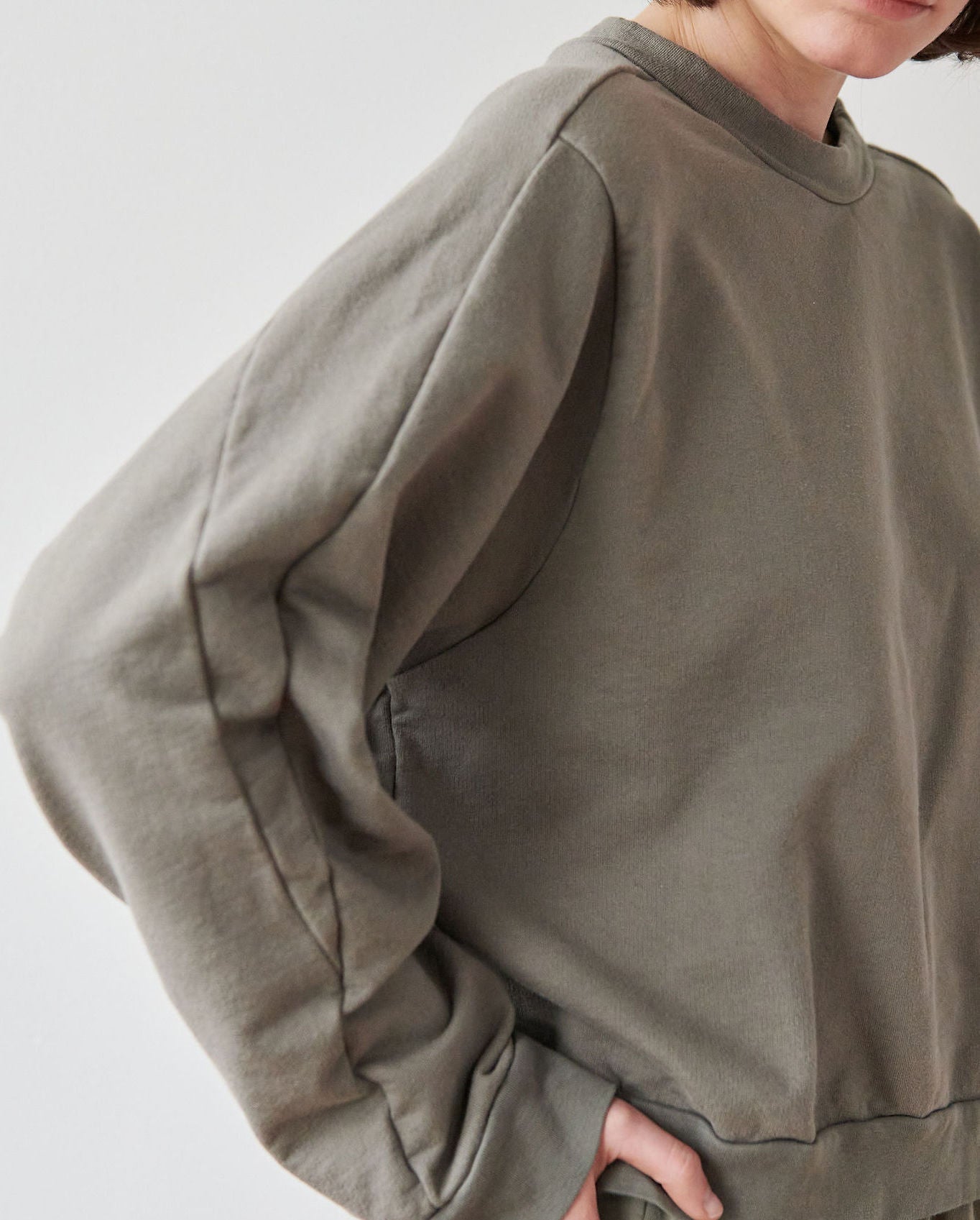 A person stands with one hand on their hip, wearing an oversized, long-sleeved, fir green Cotton French Terry Easy Summer Sweatshirt by Wol Hide. The image is cropped to show the upper body and part of the face, with the face angled slightly downwards. The background is light and out of focus.