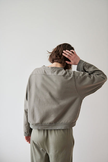 Person with short brown hair wearing the Wol Hide Cotton French Terry Easy Summer Sweatshirt in Fir and matching pants, shown from the back with one hand touching their head. The background is plain and light-colored. Made from organic cotton French terry in the USA.