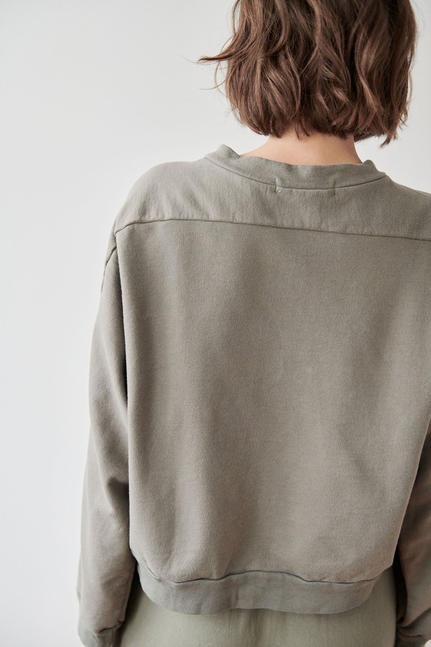 A person with shoulder-length, wavy brown hair is shown from the back wearing a loose-fitting, long-sleeved Cotton French Terry Easy Summer Sweatshirt in Fir. Made in the USA with Organic Cotton French Terry by Wol Hide, the background is plain and light-colored.