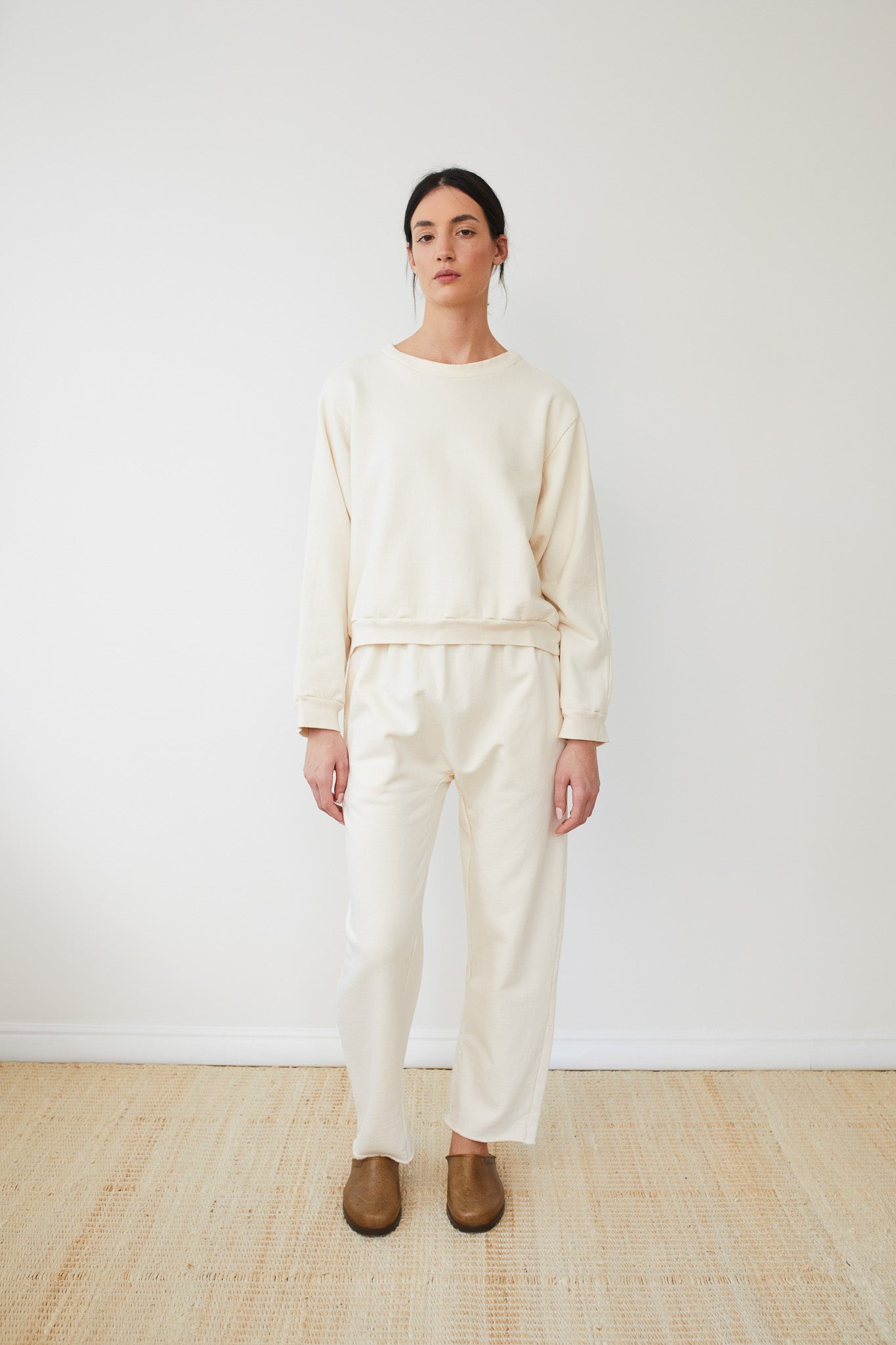 A person with dark hair is standing against a plain white background, wearing the Wol Hide Easy Winter Sweatshirt in Natural, paired with matching pants, both crafted from 100% organic cotton fleece. Complemented by brown slip-on shoes, they look directly at the camera with a neutral expression.