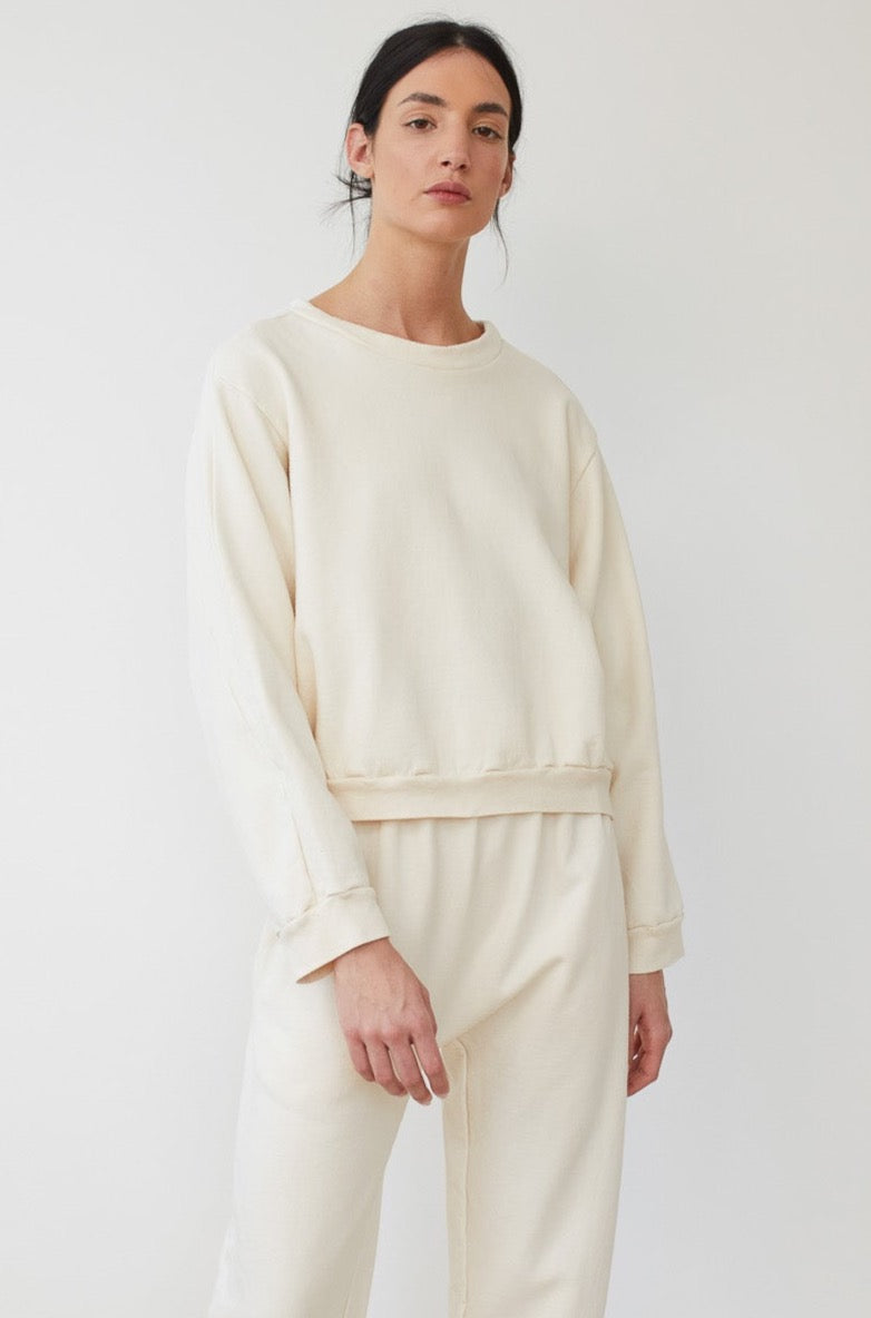 A person with long dark hair pulled back is wearing the Easy Winter Sweatshirt in Natural by Wol Hide, along with matching sweatpants. Both items are off-white, loose-fitting, and made from 100% organic cotton. They are standing against a plain light background, looking at the camera with a neutral expression.