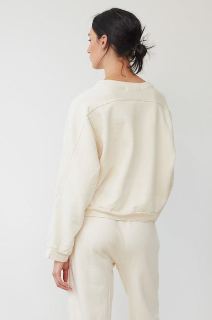 A person with dark hair in a low bun is standing with their back to the camera, wearing the Wol Hide Easy Winter Sweatshirt in Natural along with matching pants, both made in the USA from 100% organic cotton fleece. The background is plain and light-colored.