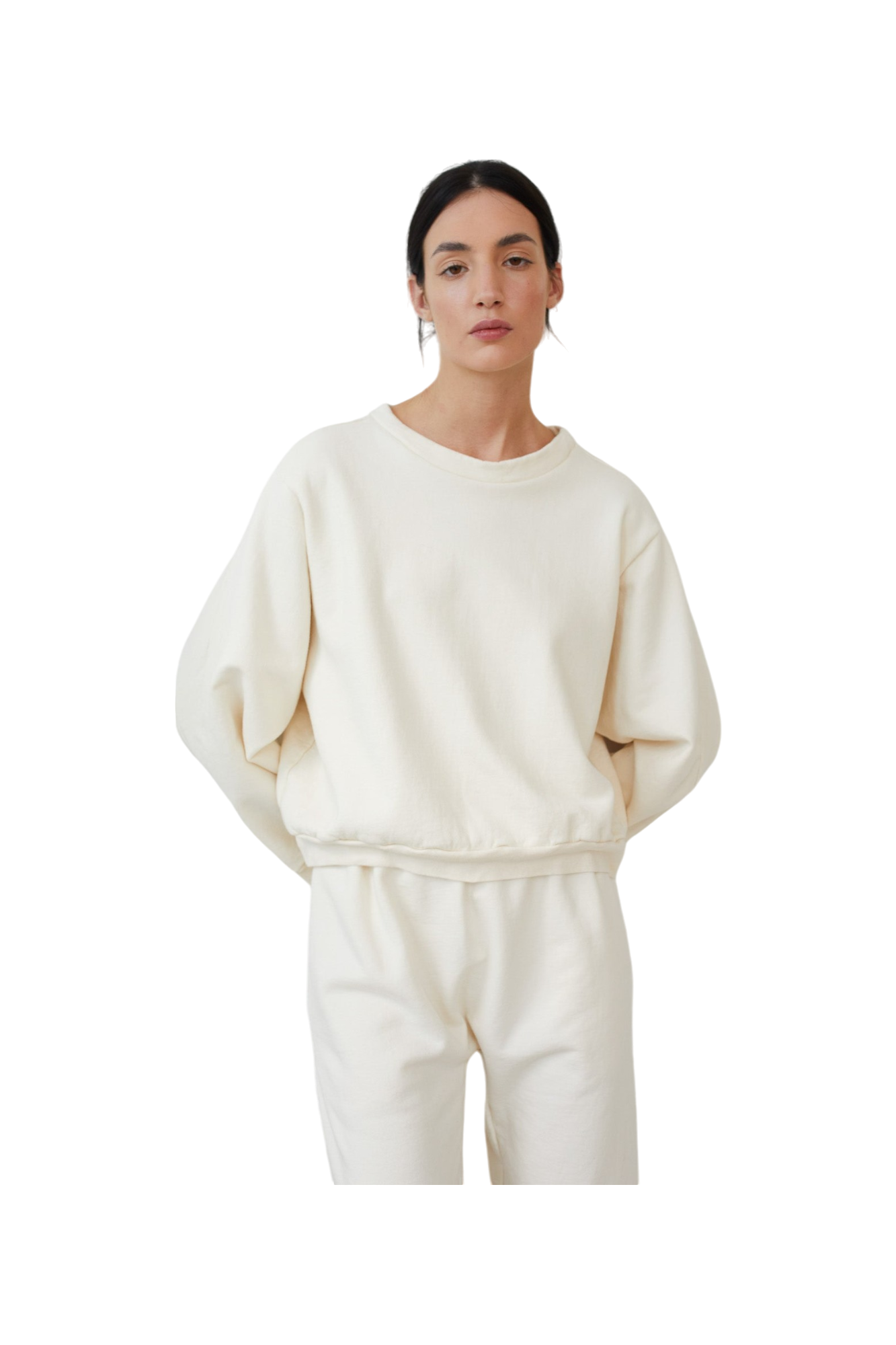 A woman with dark hair pulled back stands with a neutral expression against a plain background, wearing the Easy Winter Sweatshirt: Natural by Wol Hide. This is a cream-colored, USA-made cropped sweatshirt paired with matching pants in organic cotton fleece.