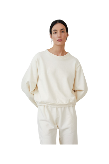 A woman with dark hair pulled back stands with a neutral expression against a plain background, wearing the Easy Winter Sweatshirt: Natural by Wol Hide. This is a cream-colored, USA-made cropped sweatshirt paired with matching pants in organic cotton fleece.