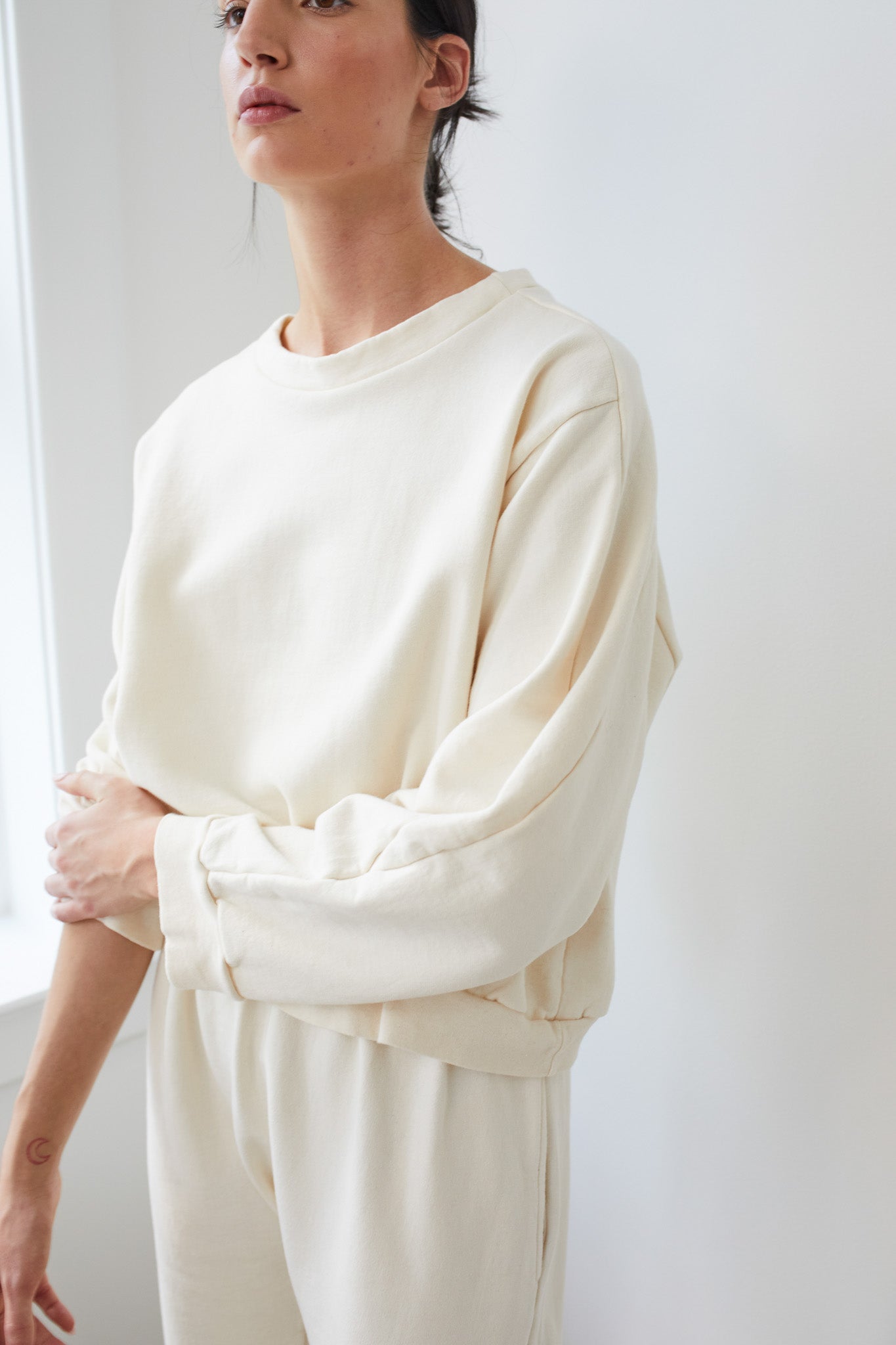 A person is wearing a soft, oversized Easy Winter Sweatshirt in natural color from Wol Hide, made of 100% organic cotton fleece, along with matching sweatpants. They are standing indoors near a window with soft natural light. Their head is slightly turned to the side, and one arm is crossed gently over the other.