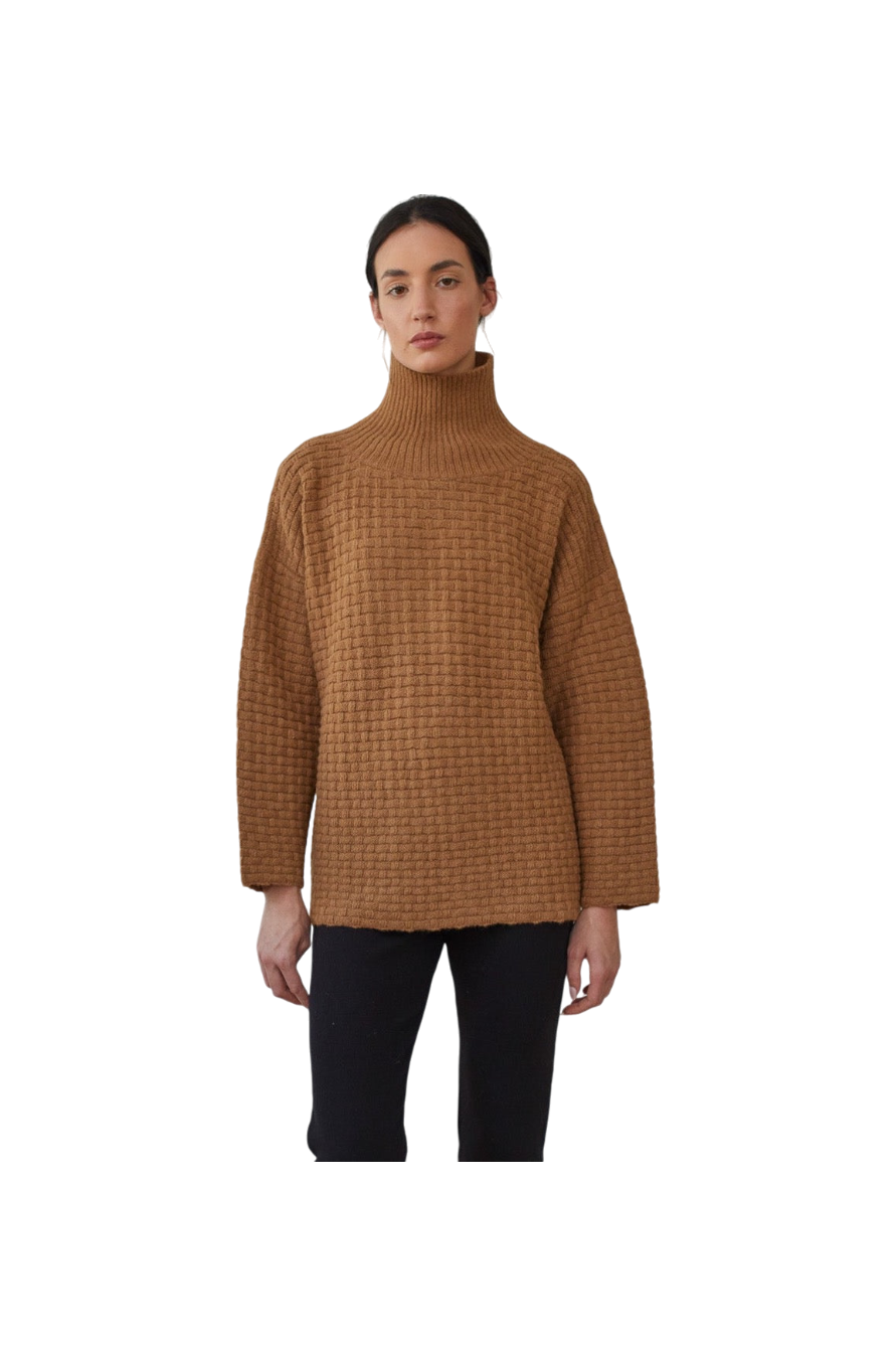 A person wearing the Wol Hide Lattice Turtleneck in Fawn, crafted with brushed alpaca yarn and paired with black pants, poses against a black background. This brown sweater showcases a basketweave textured stitch and offers a loose fit with wide sleeves.