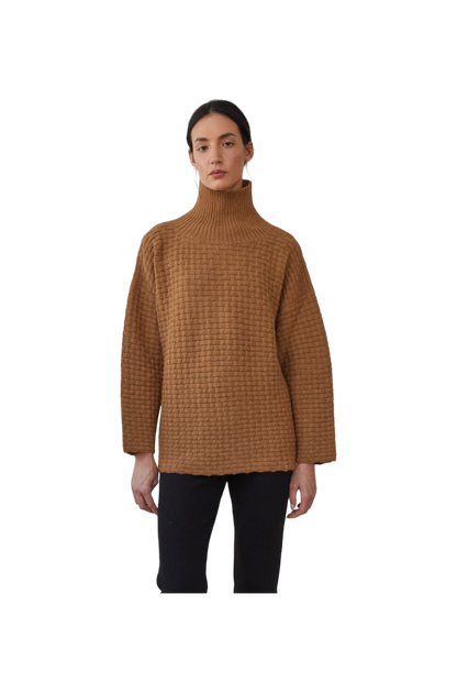 A person wearing the Wol Hide Lattice Turtleneck in Fawn, crafted with brushed alpaca yarn and paired with black pants, poses against a black background. This brown sweater showcases a basketweave textured stitch and offers a loose fit with wide sleeves.