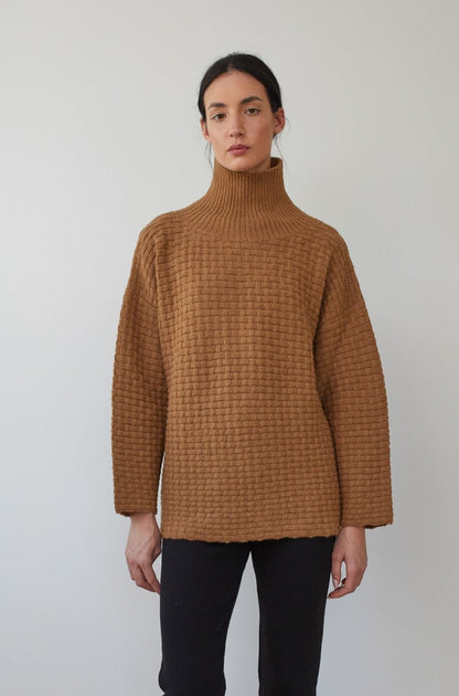 A person stands against a neutral background wearing the Wol Hide Lattice Turtleneck in Fawn, crafted with brushed alpaca yarn in a basketweave textured stitch, paired with black pants. They have a calm expression and dark hair tied back.