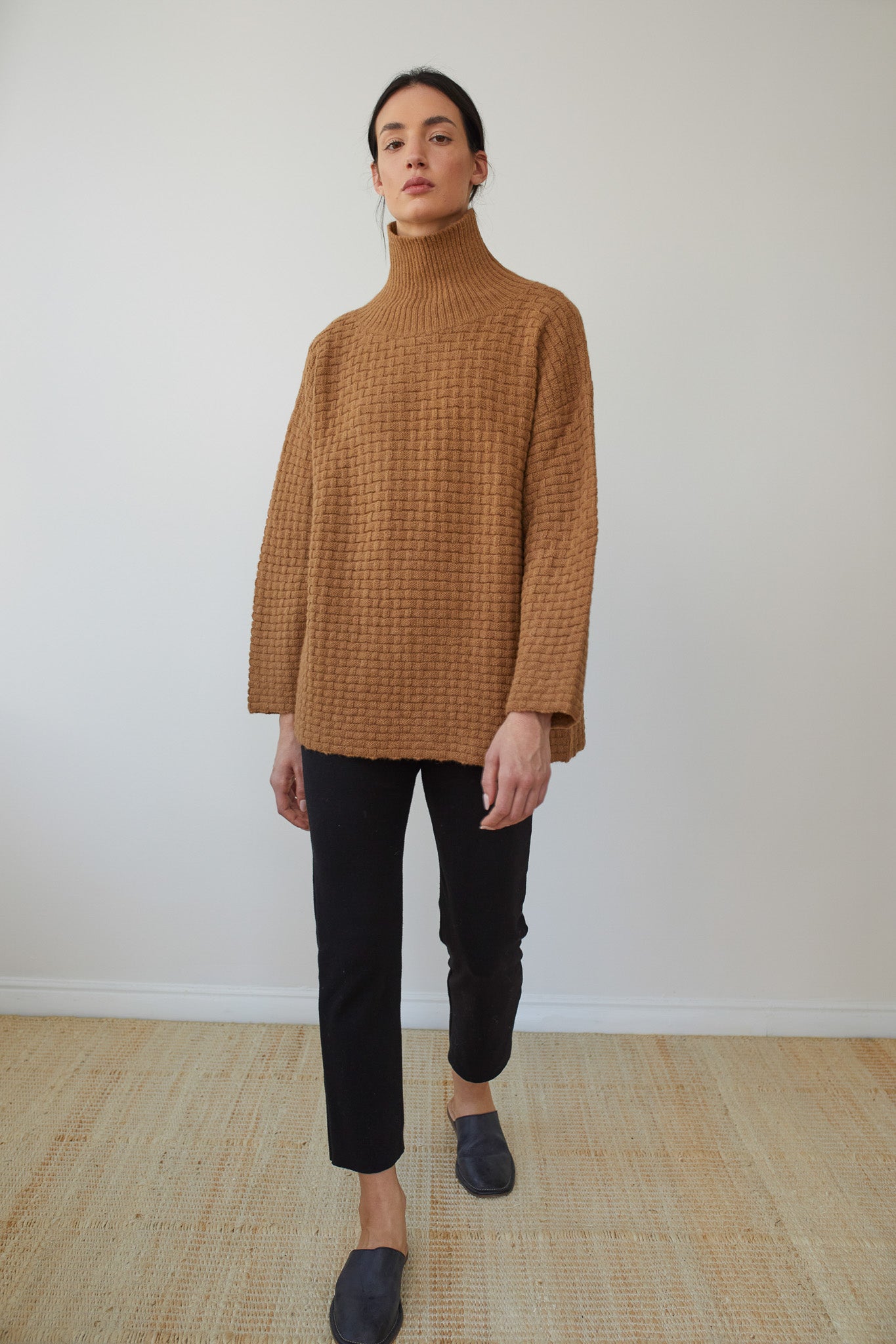 A person wearing the Lattice Turtleneck in Fawn by Wol Hide, crafted from brushed alpaca yarn, black pants, and blue slip-on shoes stands on a beige woven rug against a plain white wall. The sweater features a basketweave textured stitch.