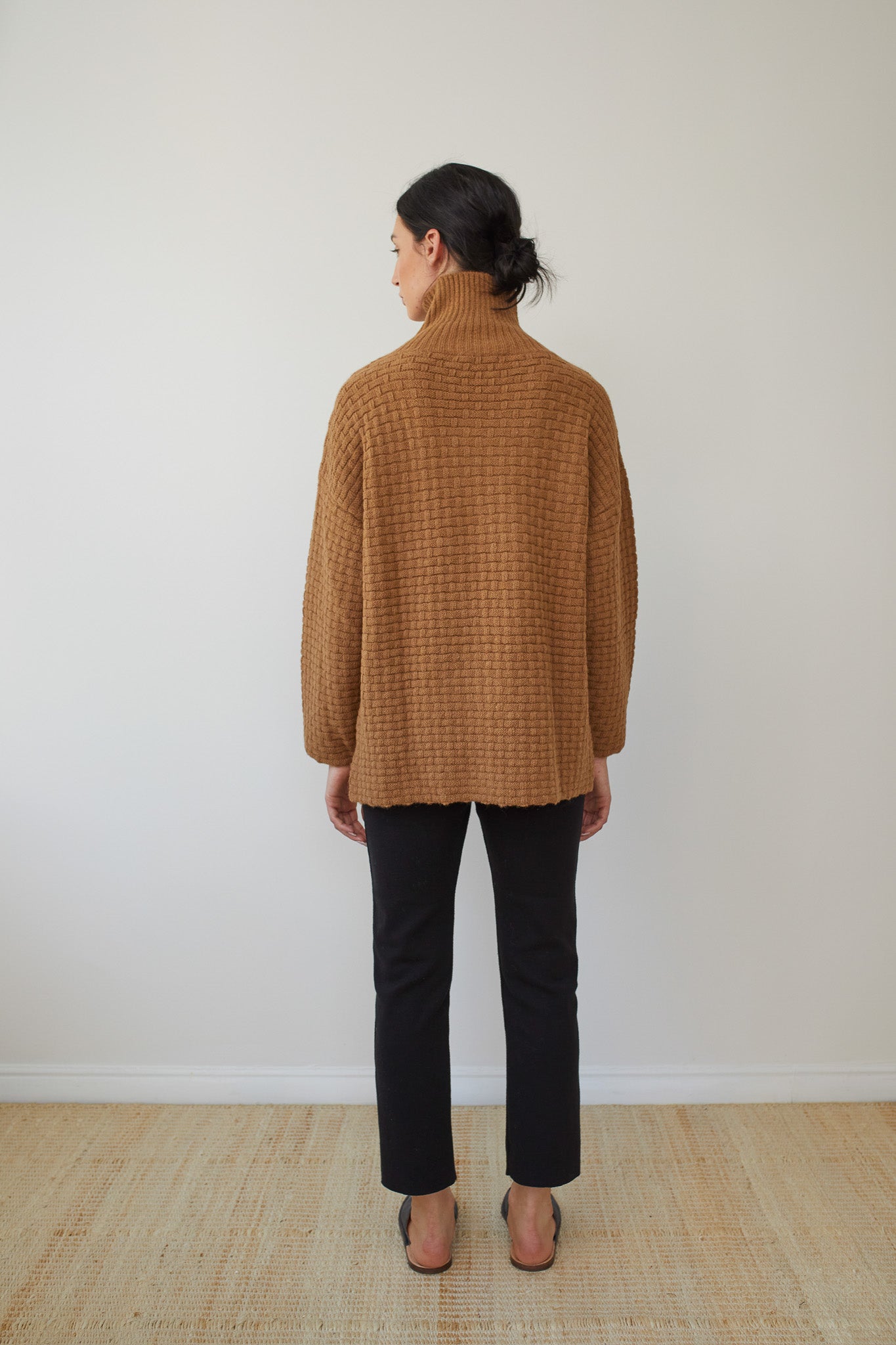 Rear view of a person with dark hair in a bun, wearing Wol Hide's Lattice Turtleneck in Fawn, featuring a basketweave textured stitch, paired with black pants. They are standing barefoot on a beige carpet against a light-colored wall.