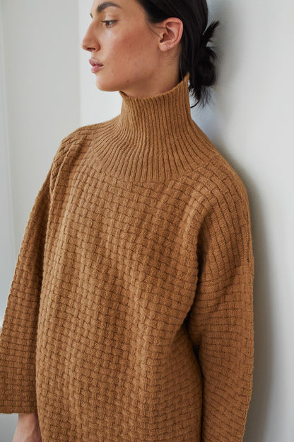 A person with dark hair in a low bun wears the Wol Hide Lattice Turtleneck in Fawn, standing against a white wall and looking to the side.