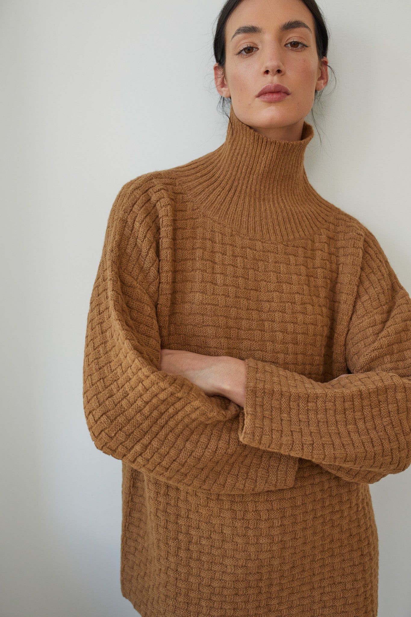 A person with dark hair tied back is wearing the Wol Hide Lattice Turtleneck in Fawn, showcasing a basketweave textured stitch, with folded arms, standing against a plain light-colored wall.