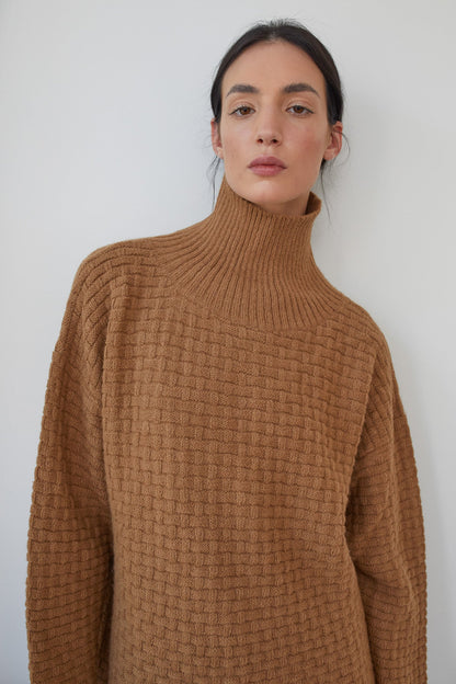 A person wearing the Lattice Turtleneck in Fawn by Wol Hide, designed with a basketweave textured stitch in brushed alpaca yarn, stands against a white background. Their hair is pulled back, and they have a neutral expression.