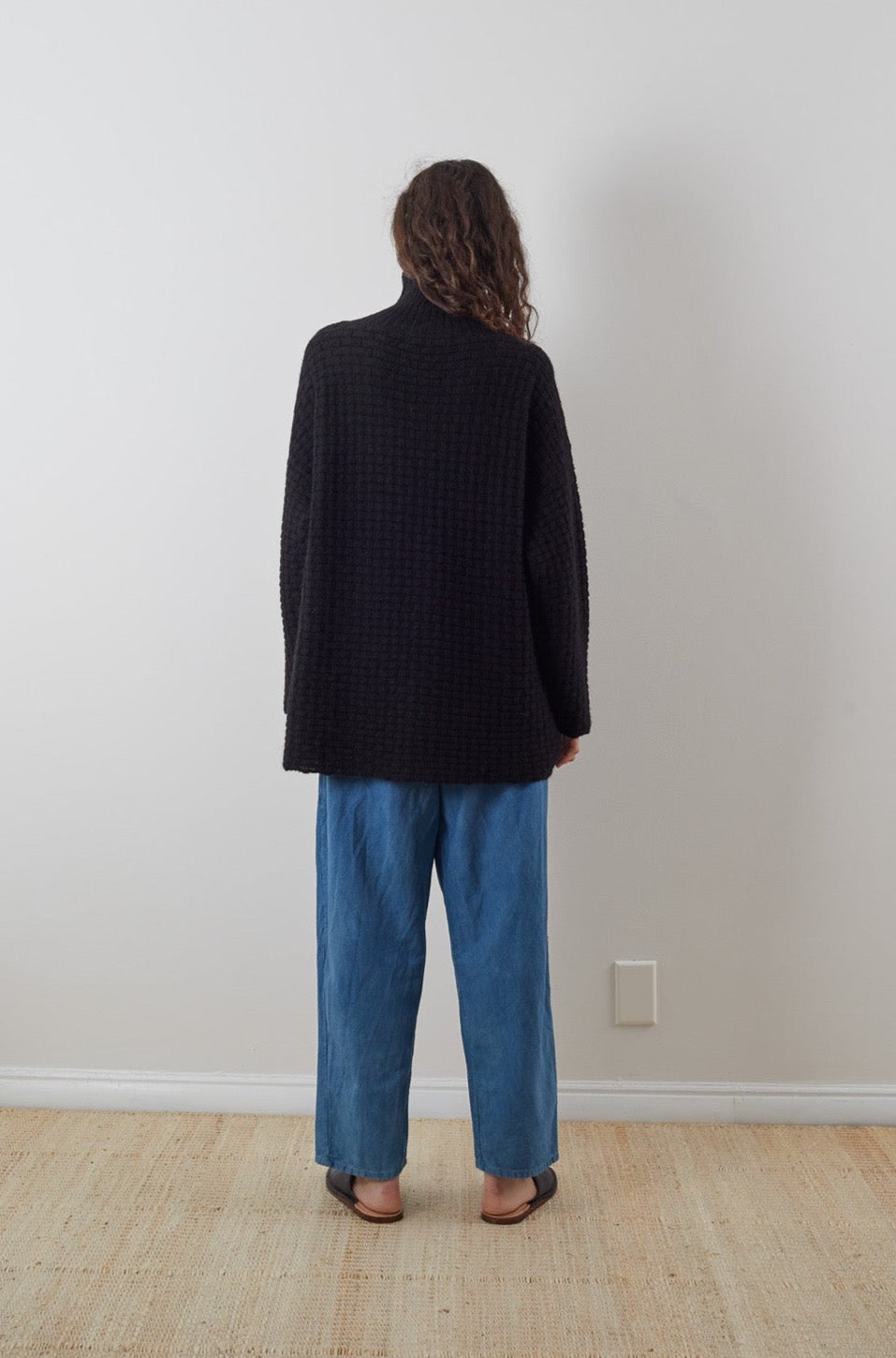 A person with long curly hair stands with their back to the camera, wearing a Wol Hide Lattice Turtleneck in Onyx. Complementing loose blue pants and sandals, they inhabit a simple indoor space, defined by its white walls and beige carpet.