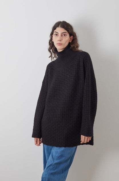 A person with long, curly hair is wearing the Lattice Turtleneck in Onyx from Wol Hide, along with loose blue pants, standing against a plain white wall. Their gaze is directed to the side.