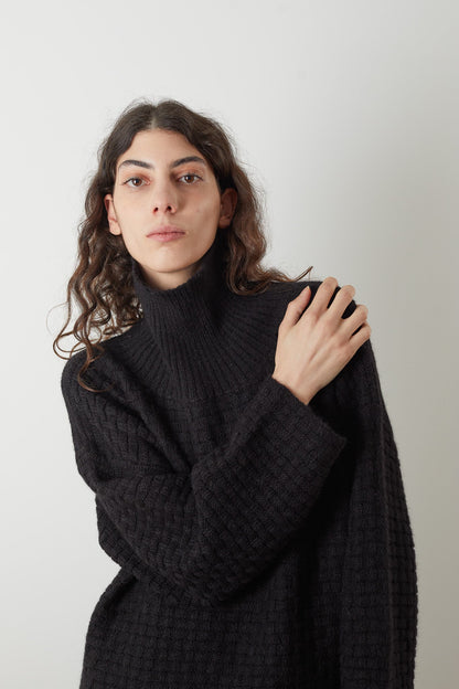 A person with long, wavy hair wears the oversized Lattice Turtleneck in Onyx by Wol Hide, crafted from brushed alpaca yarn. They have a neutral expression, with one hand resting on the opposite shoulder. The basketweave texture adds depth to the black fabric against the plain, light-colored background.