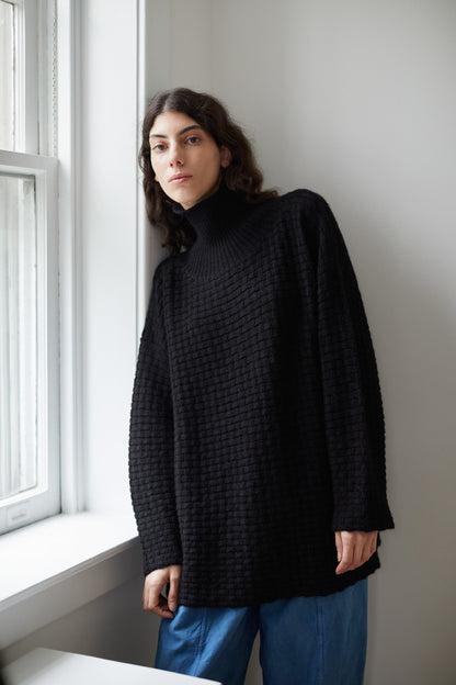 A person with long, wavy hair leans against a white windowsill, wearing the Wol Hide Lattice Turtleneck in Onyx paired with blue pants. The room's white walls and window create a calm, natural light setting.