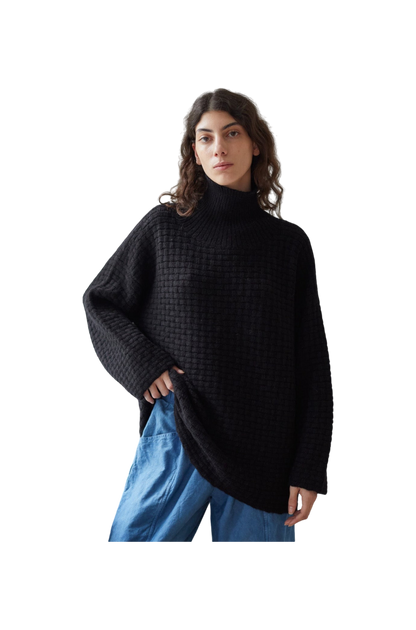 A person with long, wavy hair dons the Wol Hide Lattice Turtleneck in Onyx, crafted from brushed alpaca yarn, paired with loose blue pants. They stand against a plain background.