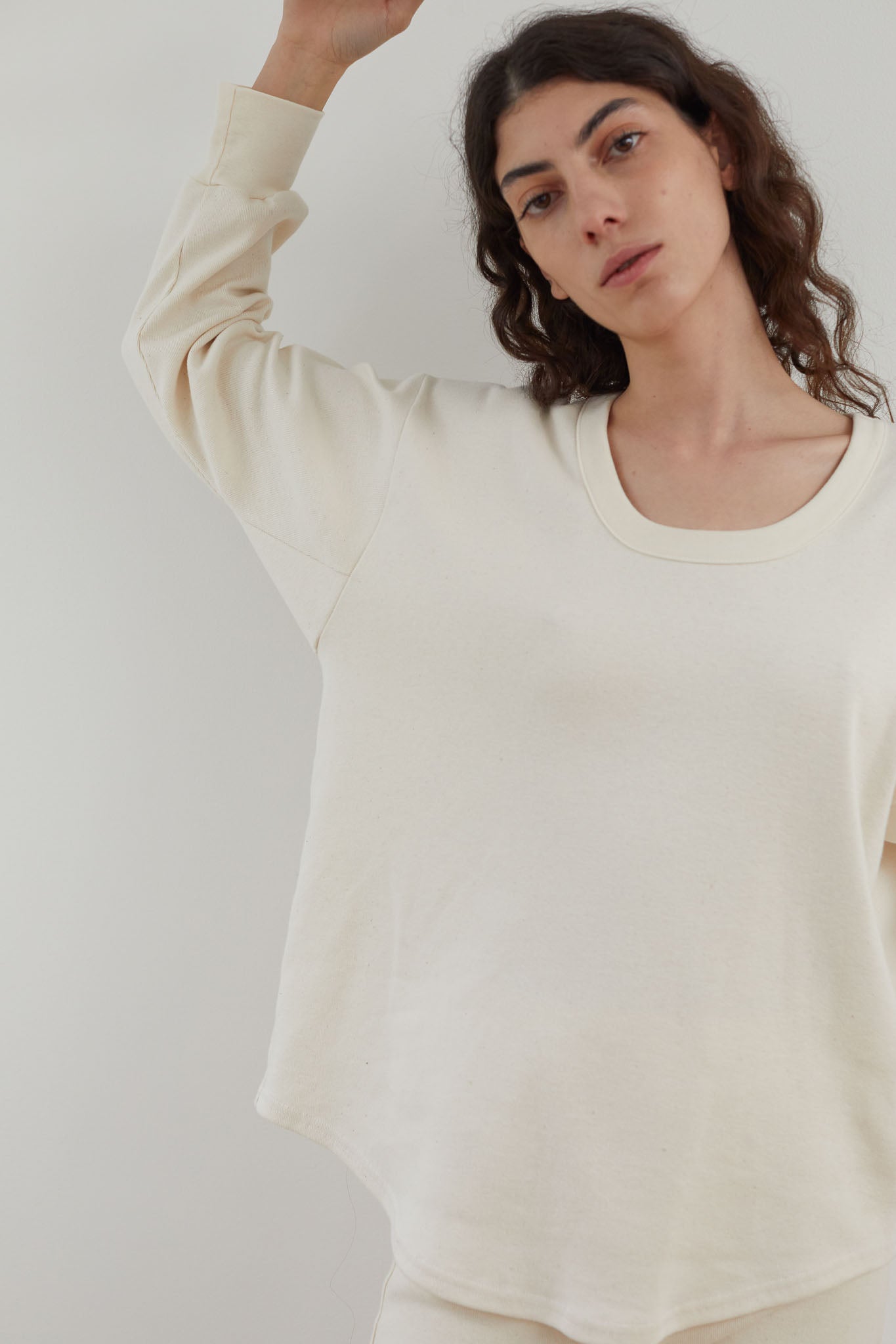 A person with long, wavy hair wears a light cream-colored Scoop Neck Thermal in Natural from Wol Hide, standing against a plain background. They have a serene expression and one arm raised slightly, embodying the simplicity and comfort of USA Made Clothing.