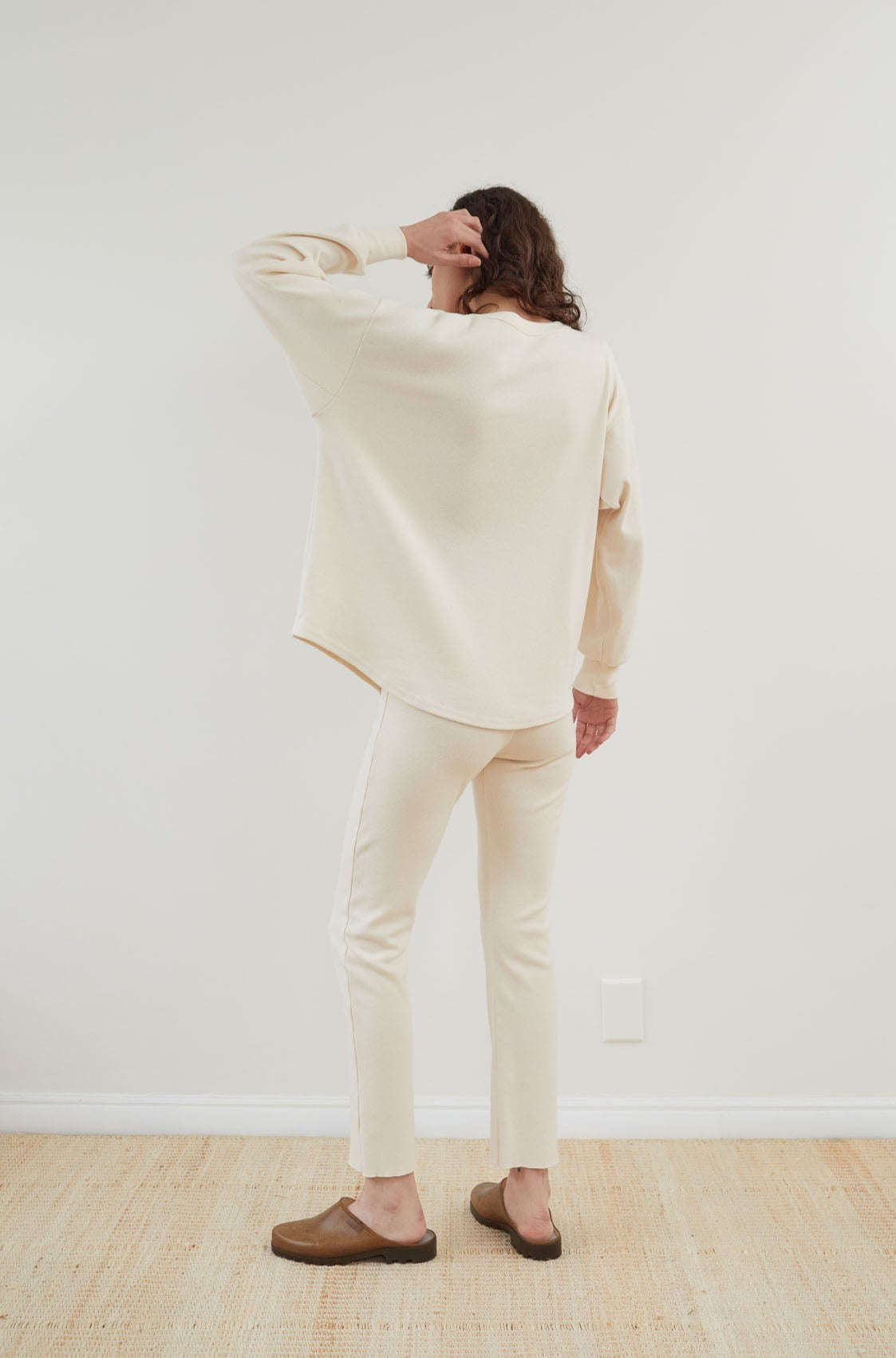 A person with long hair is standing in a minimalistic room, facing away from the camera. They are wearing Wol Hide's Scoop Neck Thermal in Natural and pants, paired with brown flat shoes. One hand is touching their hair.