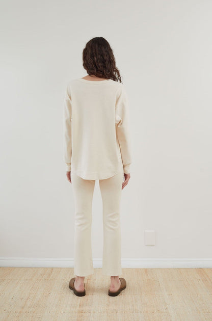 A person with long hair stands facing a white wall, wearing the Wol Hide Scoop Neck Thermal in Natural along with matching pants. They are also wearing brown shoes, standing on a light wooden floor.