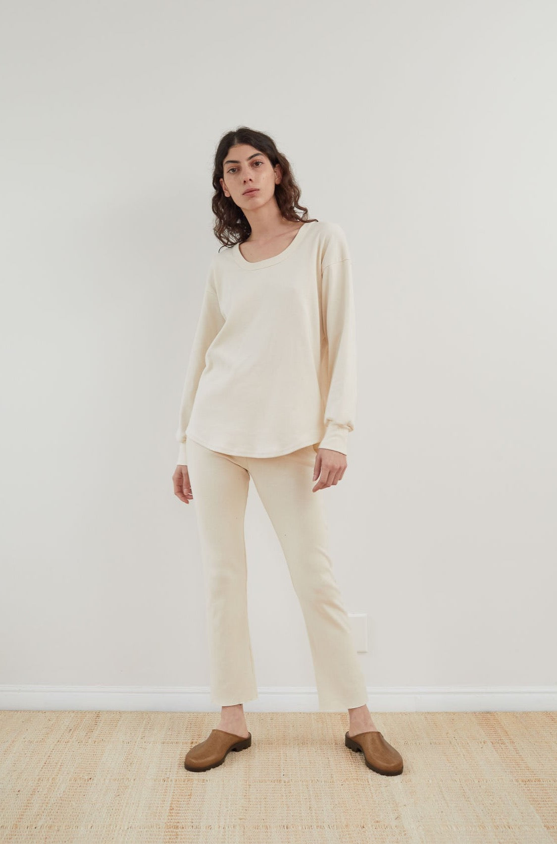 A person stands against a plain light-colored wall, wearing the Wol Hide Scoop Neck Thermal in Natural combined with matching pants. They have dark hair and are wearing brown shoes, posing on a light beige floor.