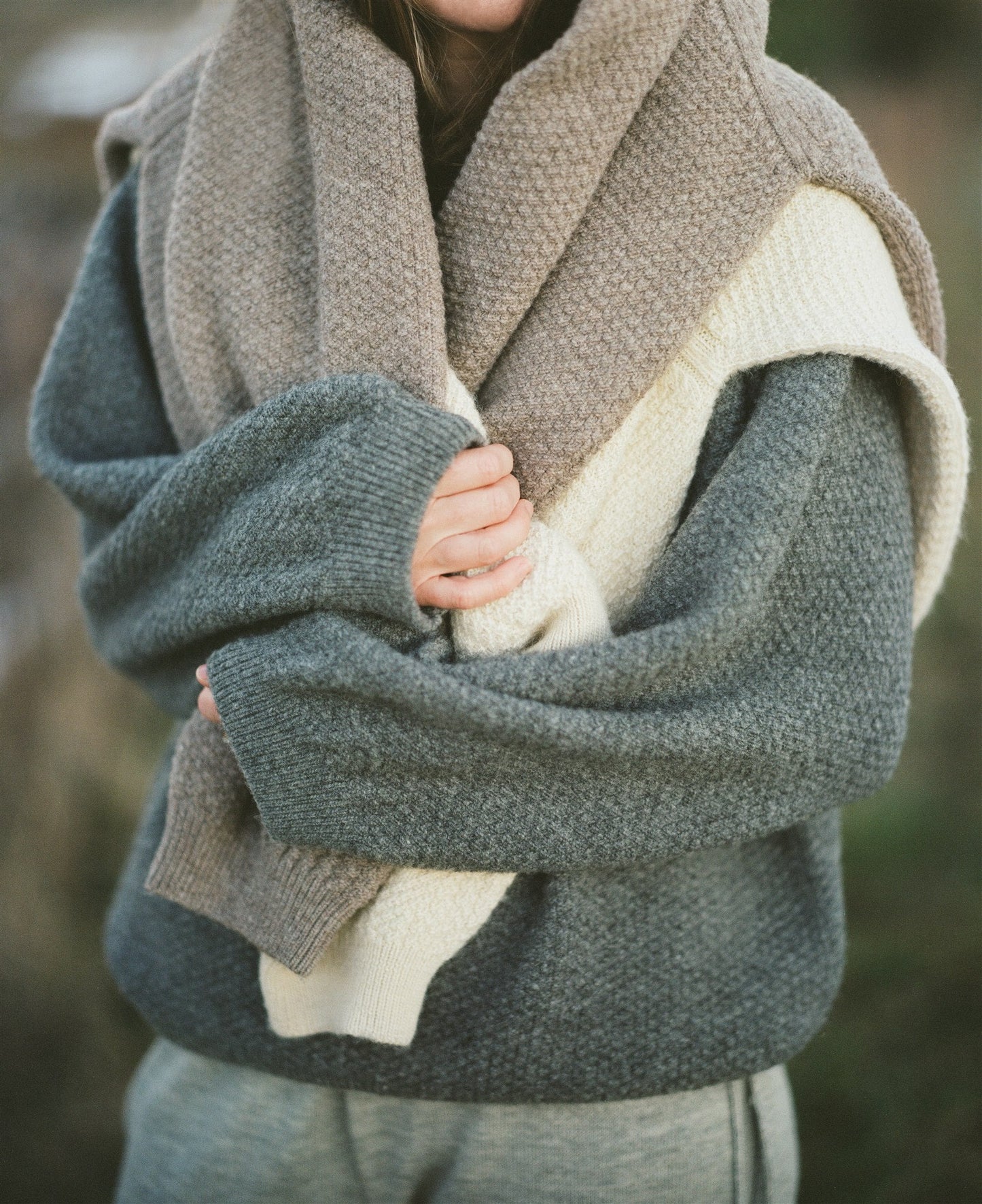 A person is wearing a cozy Earth-colored Wool and Alpaca Blend Sweatshirt Sweater by Wol Hide, holding a thick knit scarf wrapped around their shoulders. Their hands are visible, gently clasping the wool and alpaca blend scarf. The background is a blurred outdoor setting, with the person's face not visible in the image.