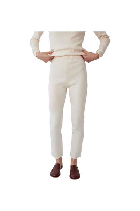 The person is dressed in a cream long-sleeve top and fitted Easy Legging: Natural from Wol Hide, with their hands on their waist. The leggings are made from stretchy rib fabric, and they complement maroon slip-on shoes. The scene is set against a plain white background.