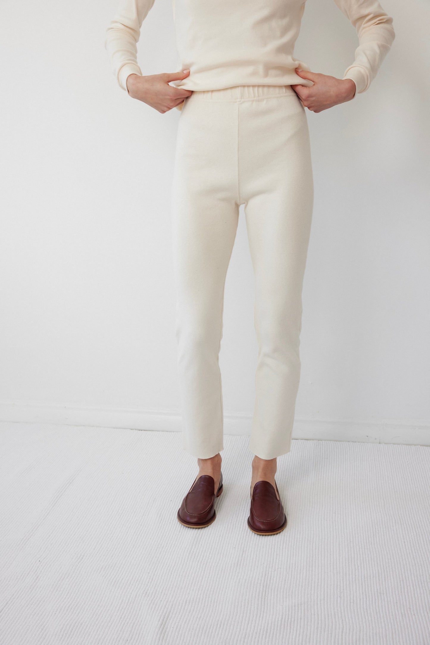 A person dressed in a cream-colored outfit with long sleeves and USA-made Easy Legging : Natural from Wol Hide, featuring an elastic waist, stands against a plain white background. They have paired the leggings with brown loafers, and their hands rest on their hips.