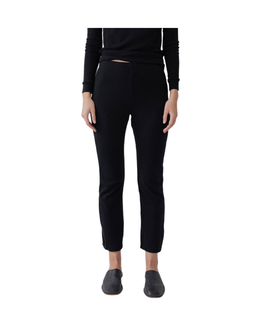 A person stands against a black background wearing Wol Hide's Easy Legging in black, made in the USA from stretchy rib fabric. The fitted pants end above the ankles and are paired with sleek slip-on shoes. They complete the look with a black long-sleeve top, arms relaxed by their sides.