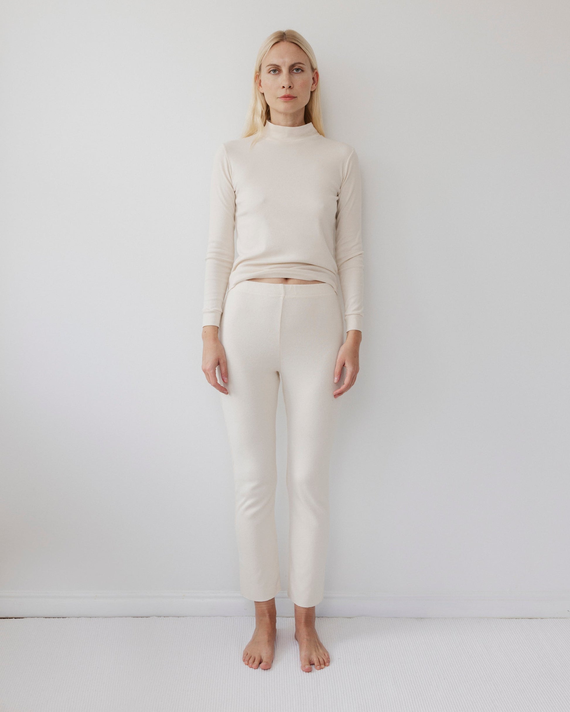 A person with long blonde hair stands against a plain white background, wearing Wol Hide's USA-made Easy Legging in Natural, crafted from cream-colored organic cotton, along with a matching heavyweight ribbed long-sleeve top. They are barefoot and maintain a neutral expression.