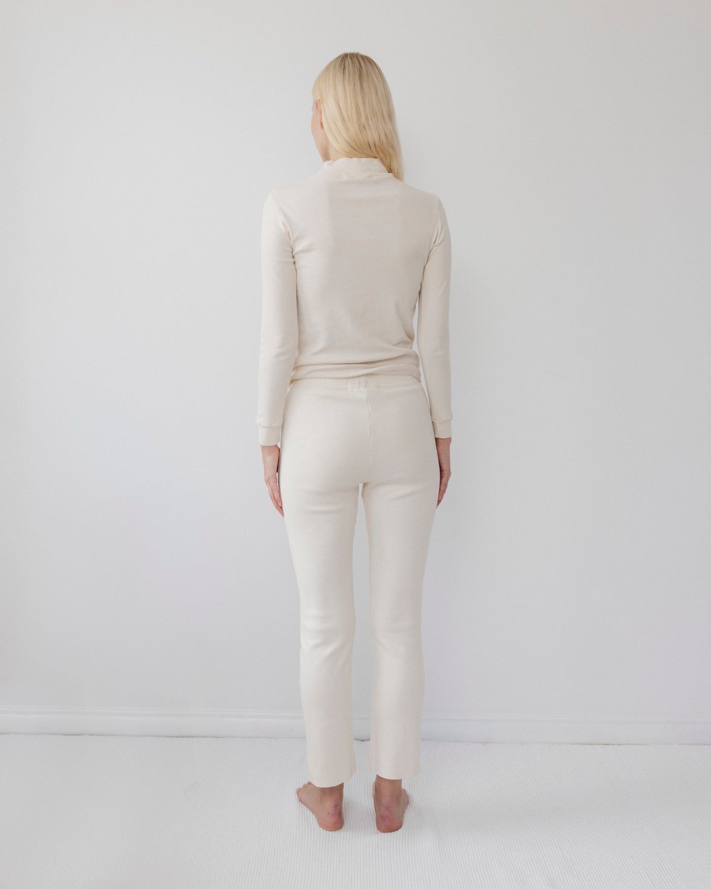 A woman with long blonde hair stands barefoot, facing away, wearing a light-colored, form-fitting ensemble featuring the Easy Legging in Natural from Wol Hide. The leggings are crafted from USA-made organic cotton and set against a plain white background.