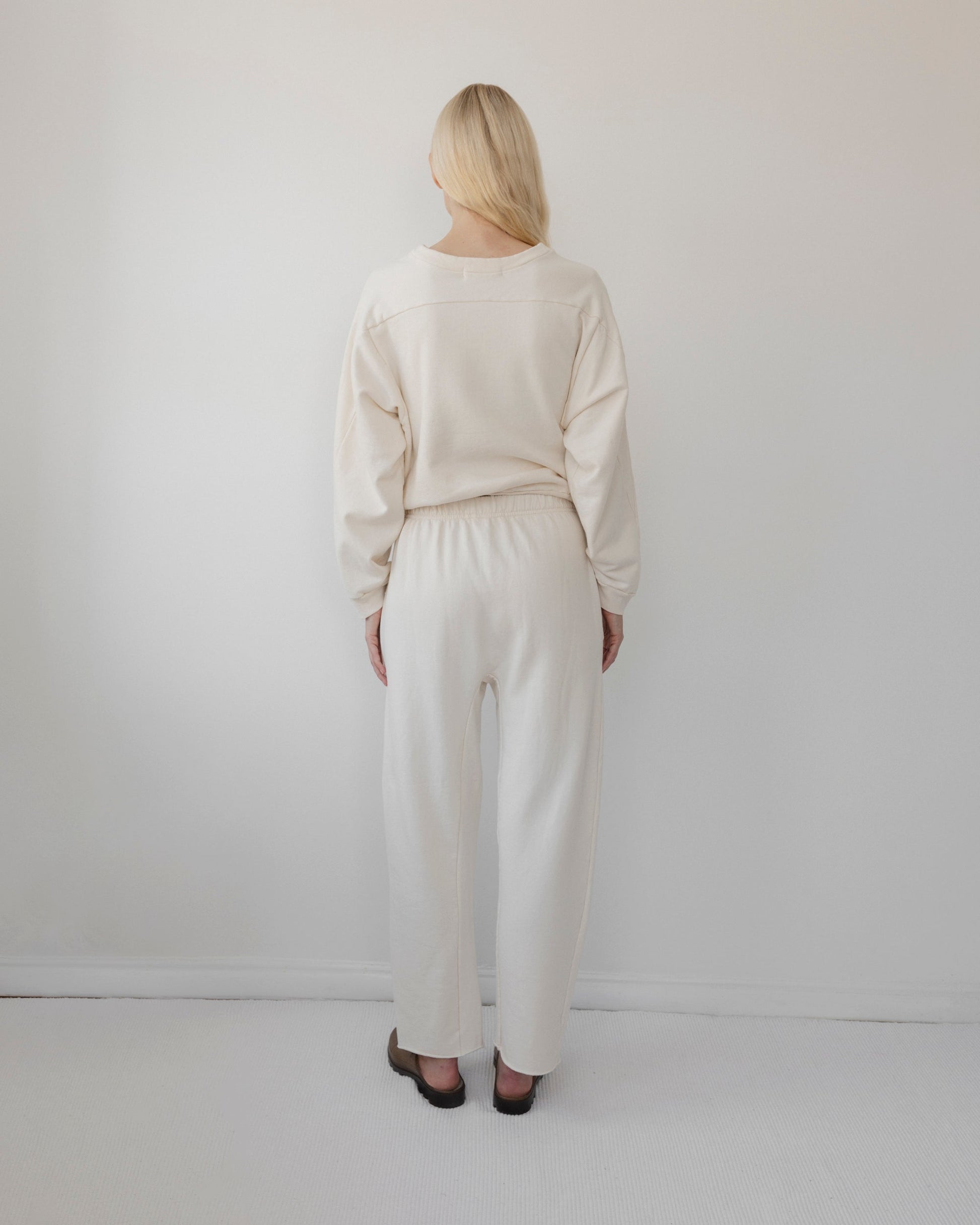 A person with long blonde hair stands facing a white wall, dressed in the Easy Sweatpant: Natural by Wol Hide and a matching cream-colored sweater. The set features an elastic waistband and is crafted in the USA from organic cotton fleece. Black sandals complete the look within this minimalistic room, which boasts a soft, neutral color palette.