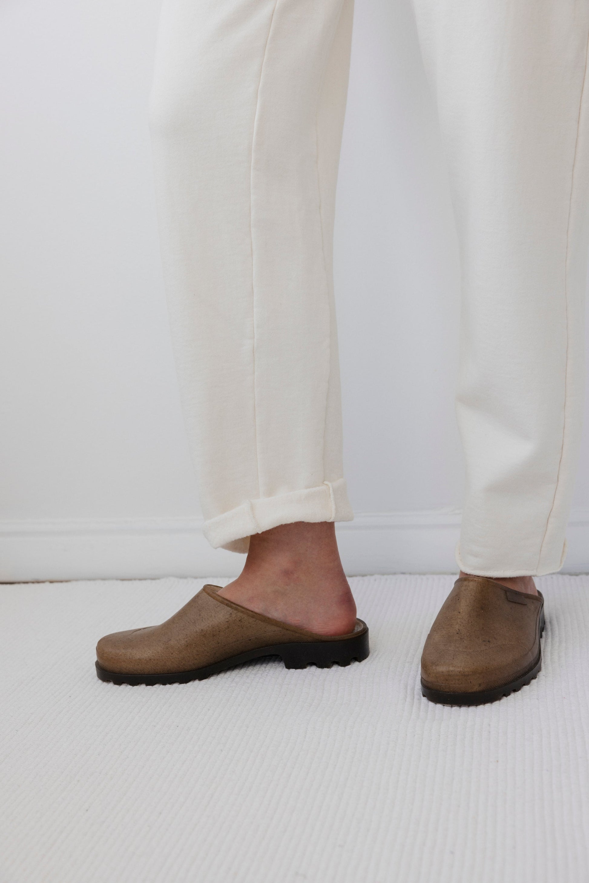 A person wearing the Natural Easy Sweatpant by Wol Hide, featuring an elastic waist, stands on a light-colored rug, showcasing brown slip-on shoes.