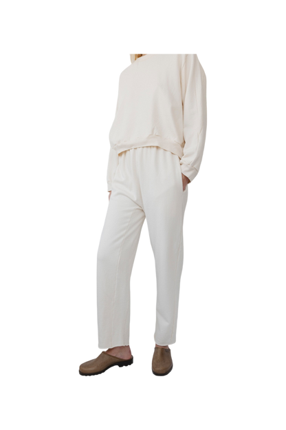 A person is wearing the Easy Sweatpant in Natural by Wol Hide, made from organic cotton fleece, consisting of a loose-fitting top and elastic waist pants, with hands in their pockets. They are also wearing brown slip-on shoes against a plain white background.