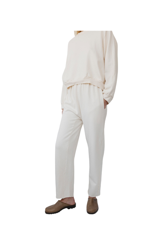 A person is wearing the Easy Sweatpant in Natural by Wol Hide, made from organic cotton fleece, consisting of a loose-fitting top and elastic waist pants, with hands in their pockets. They are also wearing brown slip-on shoes against a plain white background.