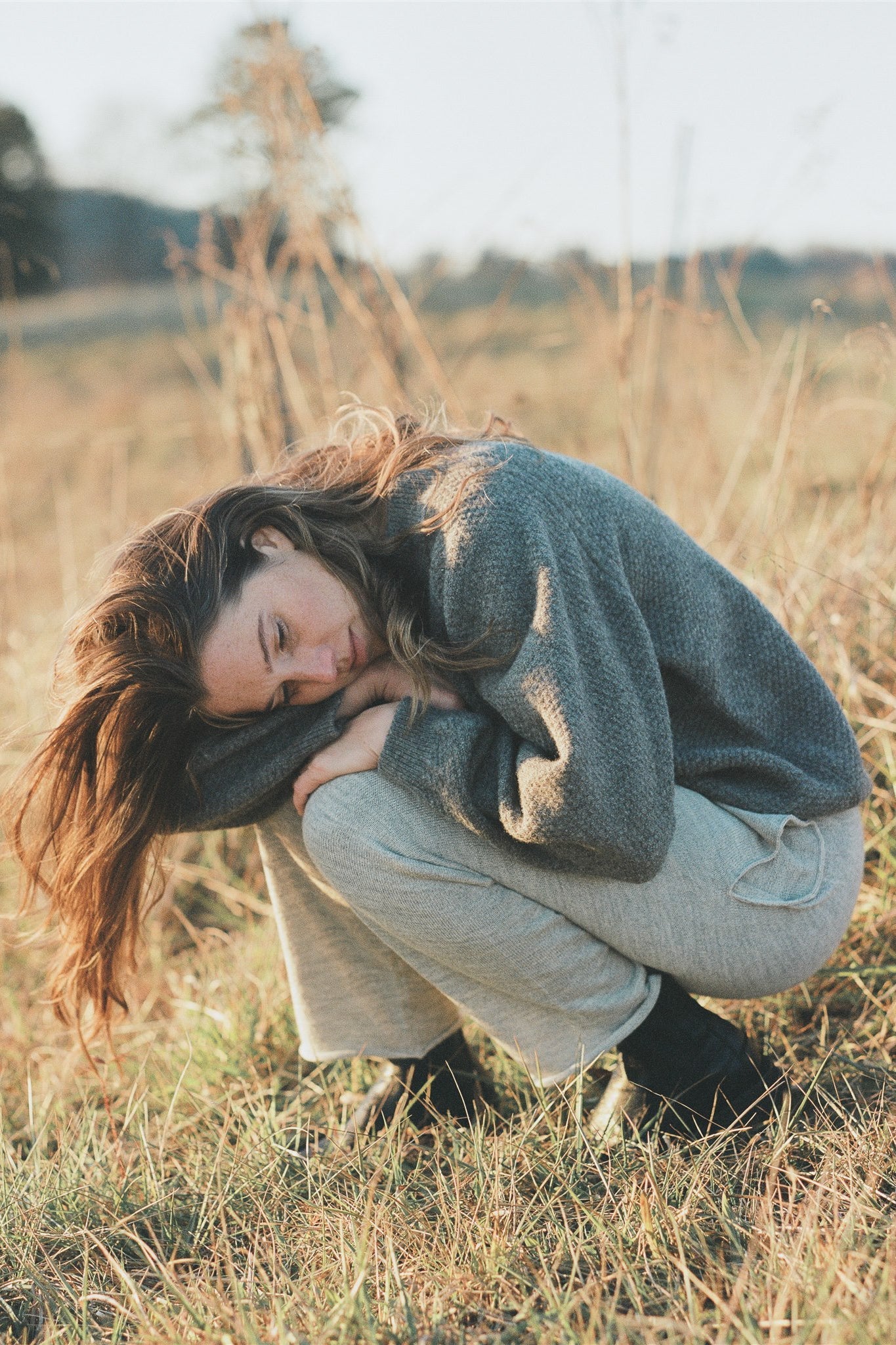 A woman with long brown hair sits in a field of tall grass, hugging her knees and looking downward. She is wearing the Wool and Alpaca Blend Sweatshirt Sweater in Earth by Wol Hide, light-colored pants, and black shoes. The lighting is warm, suggesting a sunrise or sunset, with a blurred natural background.