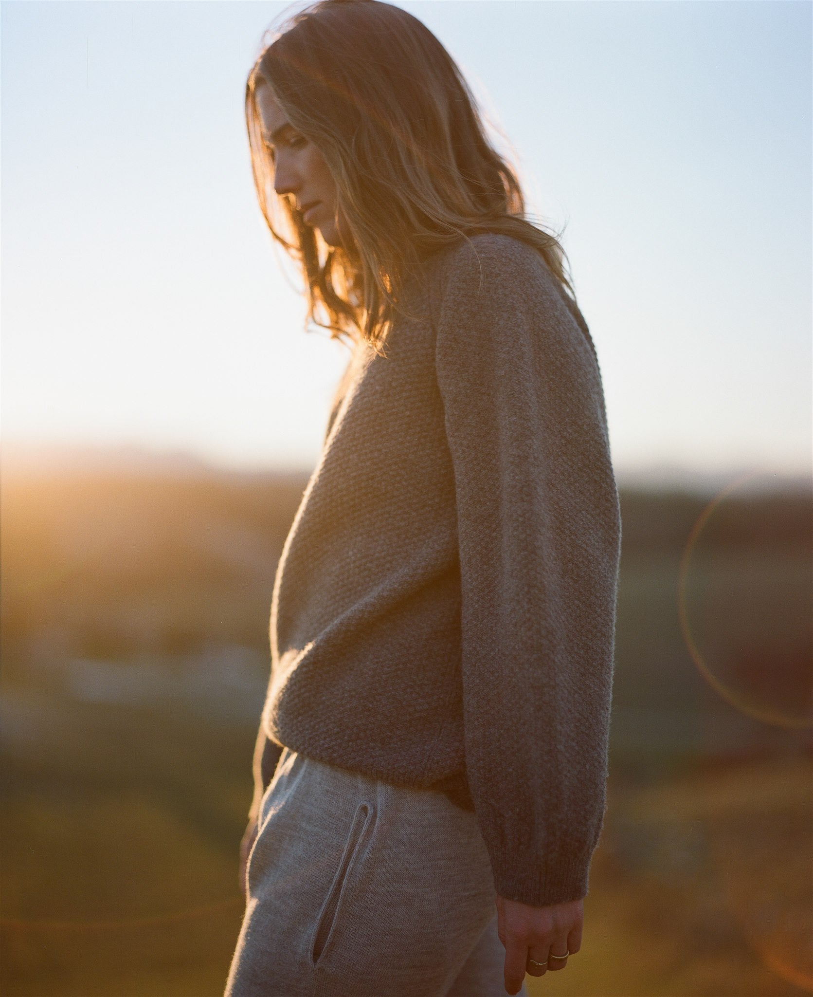 A person with long, light brown hair stands sideways at sunset, wearing the Wol Hide Wool and Alpaca Blend Sweatshirt Sweater in Earth and sweatpants. The warm sunlight casts a gentle glow, creating a serene and contemplative atmosphere.