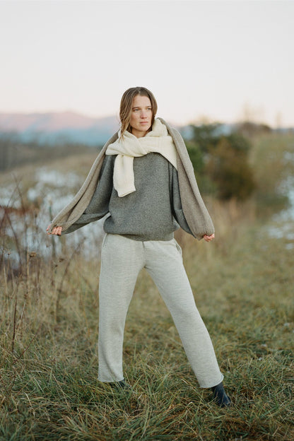 A person is posing outdoors with a distant mountain backdrop, wearing Wol Hide's Wool and Alpaca Blend Sweatshirt Sweater in Earth with raglan sleeves, light grey pants, and a cream scarf draped over their shoulders. The grassy field has a few patches of snow, and the person has a calm expression and relaxed stance.