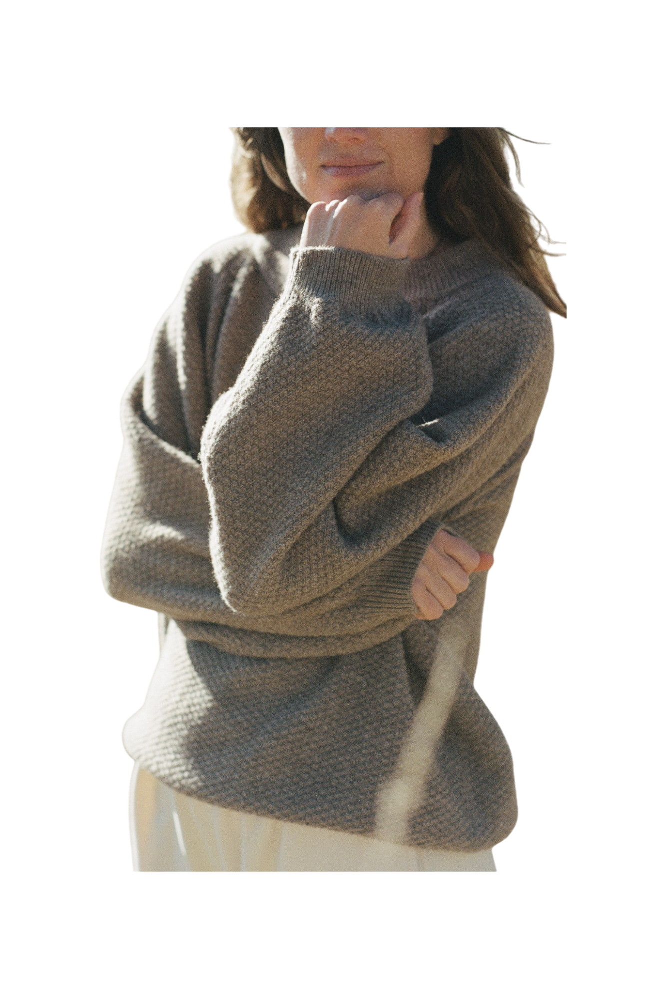 The individual, with long hair seen from the shoulders up, is wearing the Wool and Alpaca Blend Sweatshirt Sweater in Earth by Wol Hide. This chunky knit beige sweater features stylish raglan sleeves. The person has one arm crossed while the other hand rests on their chin, partially concealing their face. The background of the image is neutral. 