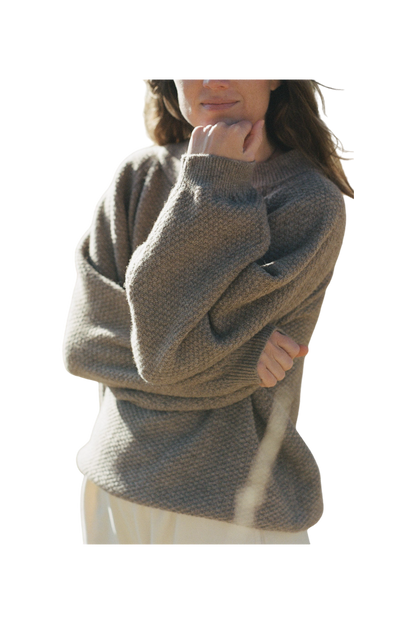 The individual, with long hair seen from the shoulders up, is wearing the Wool and Alpaca Blend Sweatshirt Sweater in Earth by Wol Hide. This chunky knit beige sweater features stylish raglan sleeves. The person has one arm crossed while the other hand rests on their chin, partially concealing their face. The background of the image is neutral. 