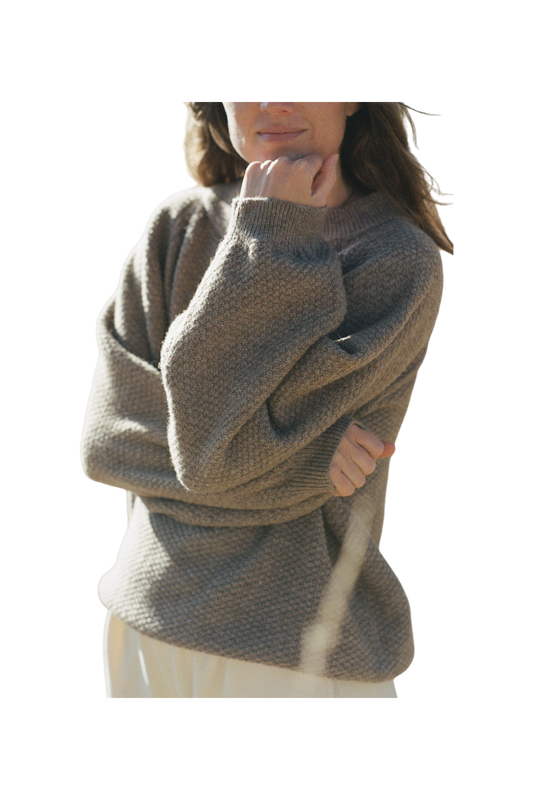 The individual, with long hair seen from the shoulders up, is wearing the Wool and Alpaca Blend Sweatshirt Sweater in Earth by Wol Hide. This chunky knit beige sweater features stylish raglan sleeves. The person has one arm crossed while the other hand rests on their chin, partially concealing their face. The background of the image is transparent.