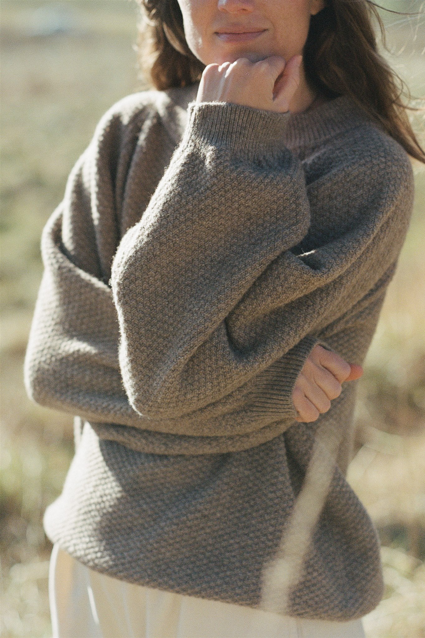 A person with long hair stands outdoors in a field, wearing the cozy, oversized Wool and Alpaca Blend Sweatshirt Sweater in Earth by Wol Hide. The person's arms are crossed, and they rest their chin on their right hand. The background is softly blurred.