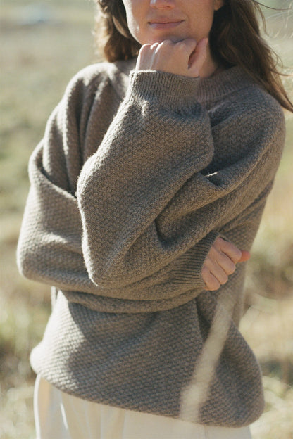 A person with long hair, dressed in a cozy Wol Hide Wool and Alpaca Blend Sweatshirt Sweater in Natural, is standing with their right hand resting on their chin and left arm crossed. They are outdoors in a grassy field, with soft natural sunlight illuminating the scene.