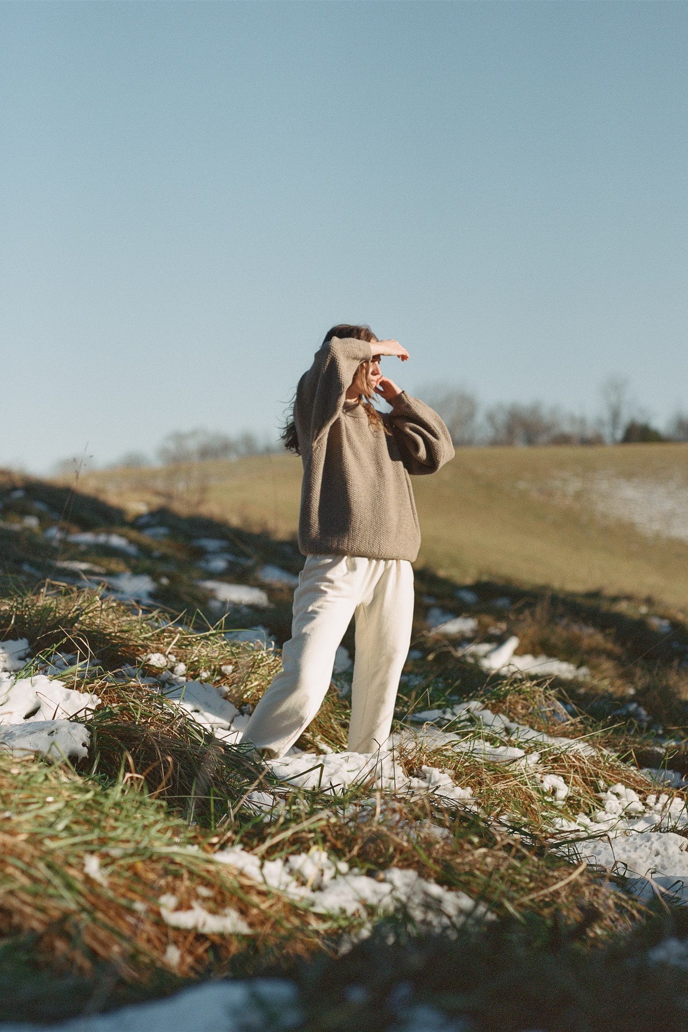 A person dressed in Wol Hide's Wool and Alpaca Blend Sweatshirt Sweater in Earth with raglan sleeves and white pants stands in a snow-dusted field, basking in the sunlight and shielding their eyes with one hand. The background features gently rolling hills and a clear blue sky.