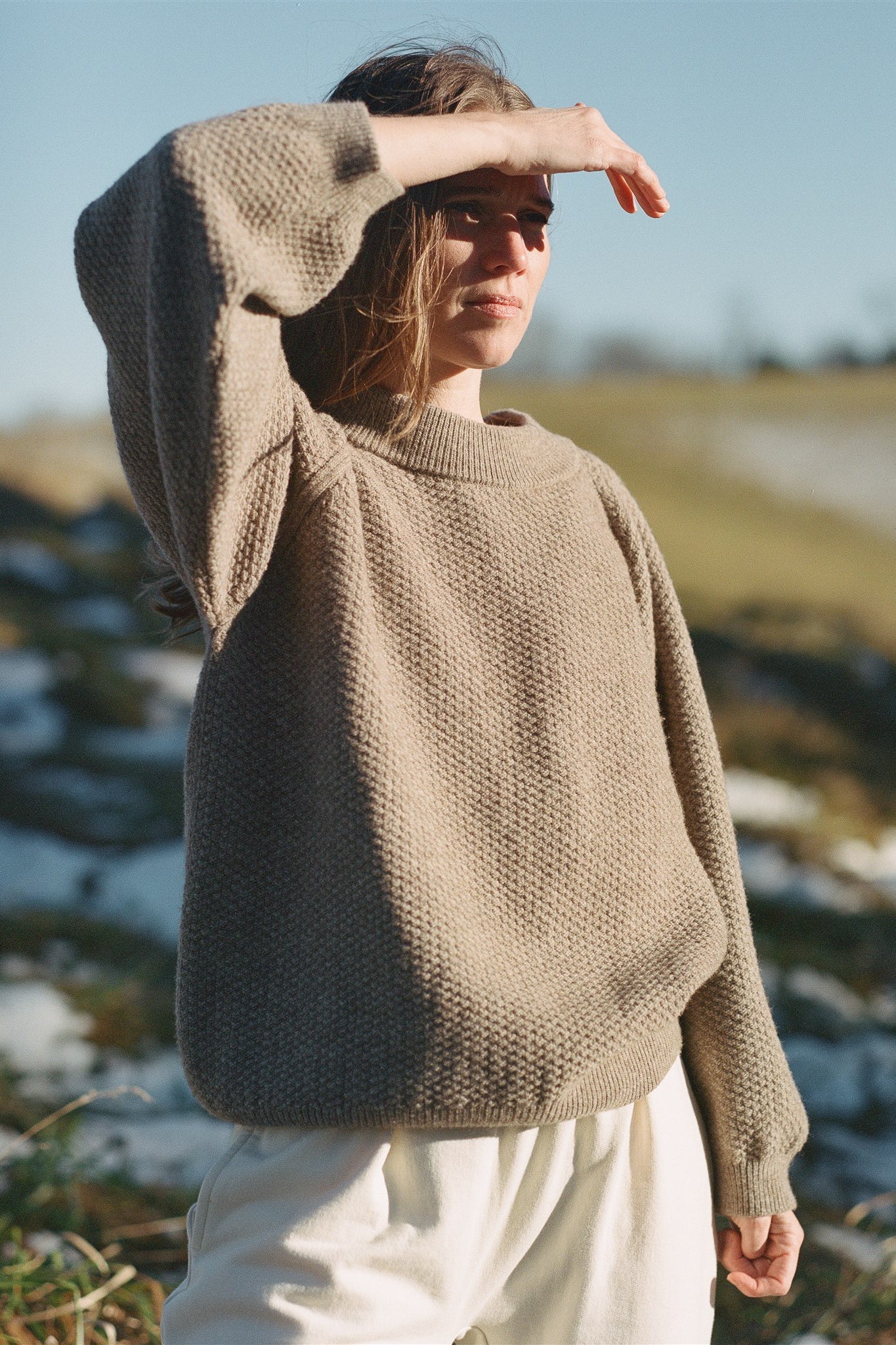 A person stands outdoors shielding their eyes from the sun with one hand. They are wearing the Wool and Alpaca Blend Sweatshirt Sweater in Earth by Wol Hide, paired with white pants. The background features grassy terrain with patches of snow under a clear blue sky.